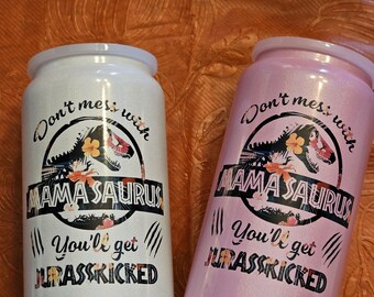 May include: Two insulated tumblers, one white and one pink, with the text "Don't mess with Mamasaurus, You'll get Jurasskicked." The design includes a dinosaur silhouette and floral accents. The tumblers have a silver rim and a black base.