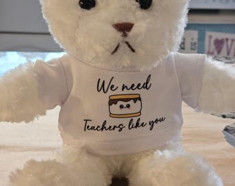 May include: White plush teddy bear wearing a white t-shirt with the text "We need teachers like you" and a cartoon image of a smiling s'more.