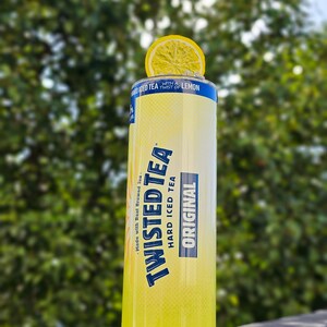 May include: A yellow can of Twisted Tea Hard Iced Tea with a lemon wedge on top. The can has blue text that reads "Made with Real Brewed Tea", "Twisted Tea", "Hard Iced Tea", and "Original". The can also has a blue stripe with the text "With a Twist of Lemon".