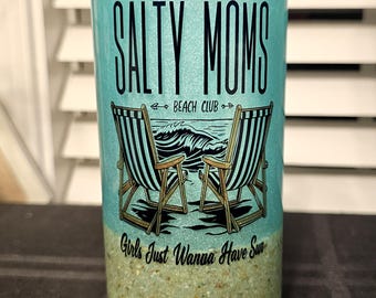 May include: A glass candle holder with a turquoise glitter design. The text "SALTY MOMS BEACH CLUB" is displayed above an illustration of two beach chairs and waves. The phrase "Girls Just Wanna Have Sun" is at the bottom.