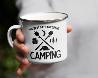 May include: White enamel camping mug with a silver rim. The mug features black graphics including crossed marshmallow roasting sticks, a tent, trees, a backpack, and the words "THE BEST DAYS ARE SPENT CAMPING."