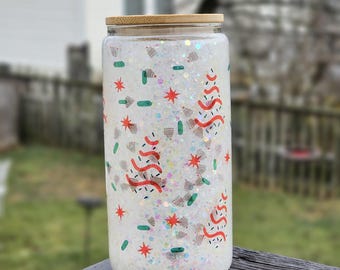 May include: A clear glass tumbler with a bamboo lid, filled with white glitter and festive decorations. The design includes red and white Christmas trees, red stars, and green sprinkles. The tumbler is sitting on a wooden surface.