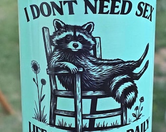 May include: A light teal tumbler with a teal lid. The tumbler features a black and white illustration of a raccoon lounging in a chair, with the text "I DON'T NEED SEX LIFE FUCKS ME DAILY."