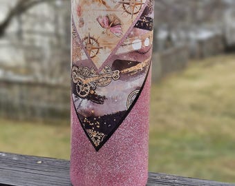 May include: A tall, insulated tumbler with a travel-themed design. The upper portion features a map and compass rose pattern with pink floral accents. The bottom half is covered in pink glitter. The tumbler has a clear lid.