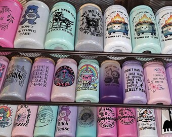 May include: A variety of glass beverage cans with colorful designs and humorous text. The cans are arranged on shelves and feature illustrations of bears, raccoons, and other graphics. The text includes phrases like "Don't Fucking Care" and "Fuck My Balls."