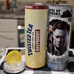 May include: Two 20 oz. insulated tumblers. The left tumbler is yellow and features a Twisted Tea logo with the words "Raspberry Hard Iced Tea" and "Made with Real Brewed Tea". The right tumbler is white and features a graphic of a character from the movie "It" with the text "House of Horrors" and "Bodies of Missing Kin Found in Derry City Sewer System".