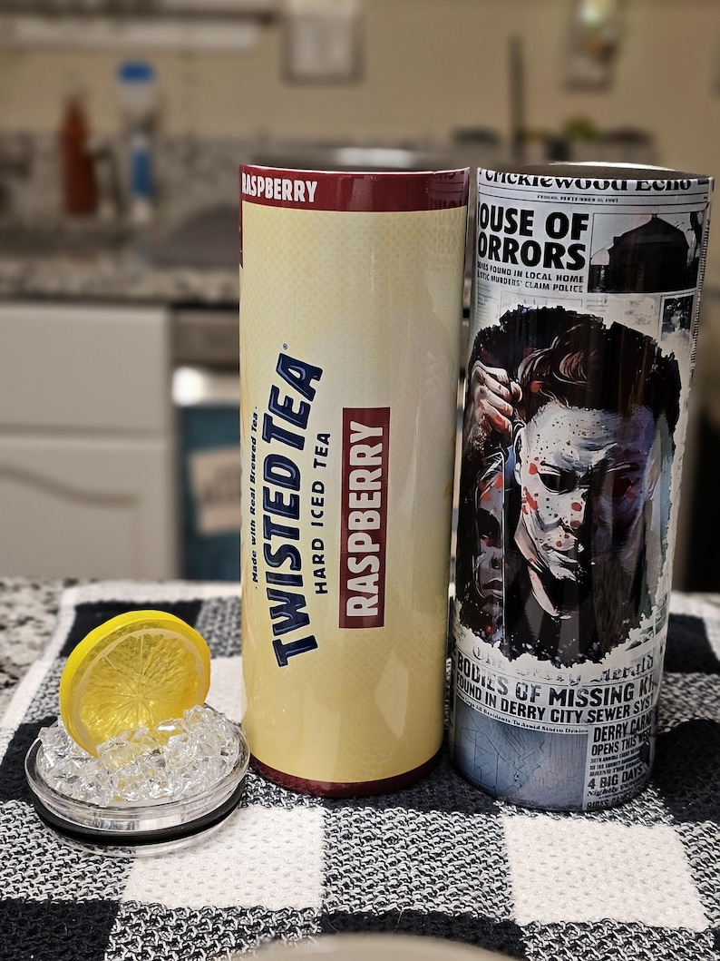 May include: Two 20 oz. insulated tumblers. The left tumbler is yellow and features a Twisted Tea logo with the words "Raspberry Hard Iced Tea" and "Made with Real Brewed Tea". The right tumbler is white and features a graphic of a character from the movie "It" with the text "House of Horrors" and "Bodies of Missing Kin Found in Derry City Sewer System".
