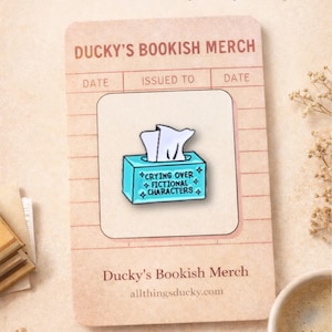 May include: Enamel pin with a blue tissue box design, featuring the text "CRYING OVER FICTIONAL CHARACTERS." The pin is on a card that reads "Ducky's Bookish Merch." The card also has the text "BOOKISH ENAMEL PIN" and "For readers who always carry a story."