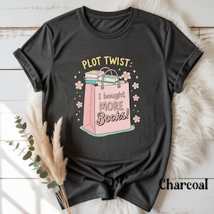 May include: Dark grey t-shirt with the slogan "PLOT TWIST: I bought MORE Books!" printed on the front. The design shows a pink shopping bag filled with books, decorated with flowers and stars. The shirt is made of a soft material.