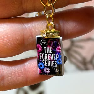 May include: A miniature book charm featuring a black cover with colorful floral accents and the white text THE FOREVER SERIES. The charm is attached to a gold-toned metal chain link with a small decorative crystal-encrusted bail at the top.