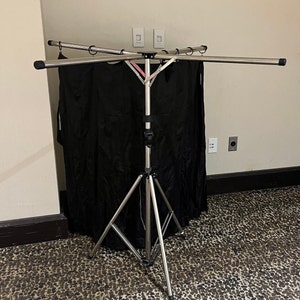 May include: A silver tripod stand with three adjustable legs and a horizontal bar with three arms. The stand is holding a black backdrop.
