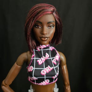 Pode incluir: Uma boneca de pele escura e olhos castanhos, com cabelo curto castanho com reflexos vermelhos. A boneca veste um top curto sem mangas preto com um padrão de grade branco e o texto rosa repetido GRL PWR, combinado com calças pretas.