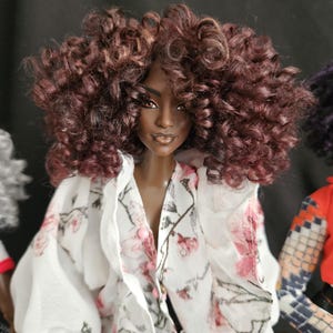 May include: A doll with dark skin and voluminous, curly auburn hair. The doll wears a white, long-sleeved garment featuring a delicate pink and black floral pattern, accented with small pink buttons down the center front of the bodice.