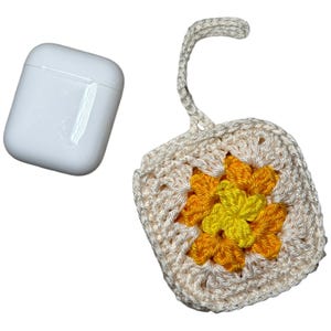 Könnte beinhalten: Ein weißes AirPod-Etui neben einer cremefarbenen, gehäkelten AirPod-Etui-Hülle. Die Hülle hat ein quadratisches Design mit einer gelb-orangen Blume in der Mitte und eine Schlaufe zum Befestigen an einem Schlüsselanhänger.