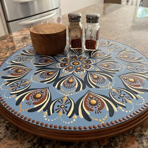 May include: A round wooden lazy susan featuring a light blue surface with a hand-painted mandala design in shades of dark blue, brown, cream, and gold. A small wooden bowl and two glass salt and pepper shakers with silver lids sit on top.