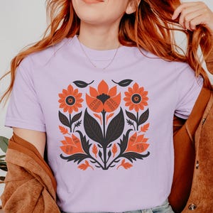 May include: A light purple t-shirt featuring a floral design in orange, black, and white. The graphic includes stylized flowers, leaves, and other botanical elements. The shirt is made of a soft material.