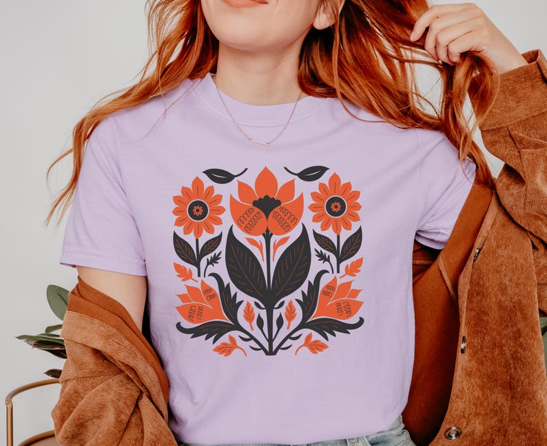 May include: A light purple t-shirt featuring a floral design in orange, black, and white. The graphic includes stylized flowers, leaves, and other botanical elements. The shirt is made of a soft material.