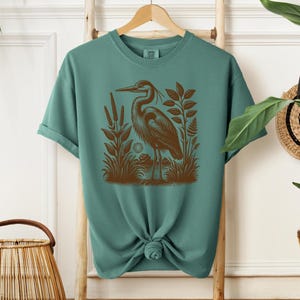 May include: Teal blue t-shirt with a brown graphic print of a heron standing in a marsh.