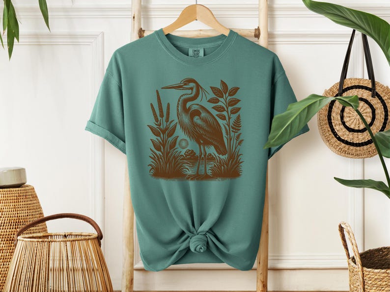 May include: Teal blue t-shirt with a brown graphic print of a heron standing in a marsh.