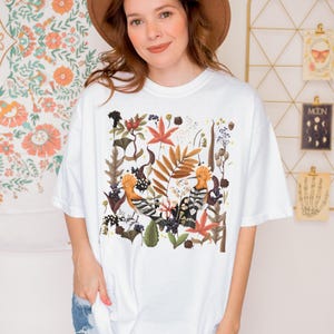 May include: White t-shirt with a colorful illustration of two birds surrounded by leaves and flowers. The birds are orange, black, and white. The leaves and flowers are in shades of green, brown, orange, and red.