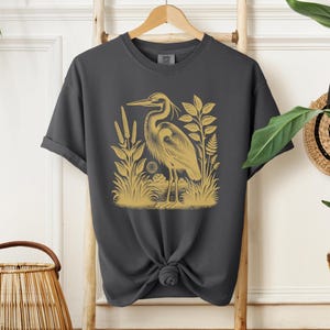 May include: A dark gray t-shirt with a gold illustration of a heron standing in a marsh with reeds and plants.