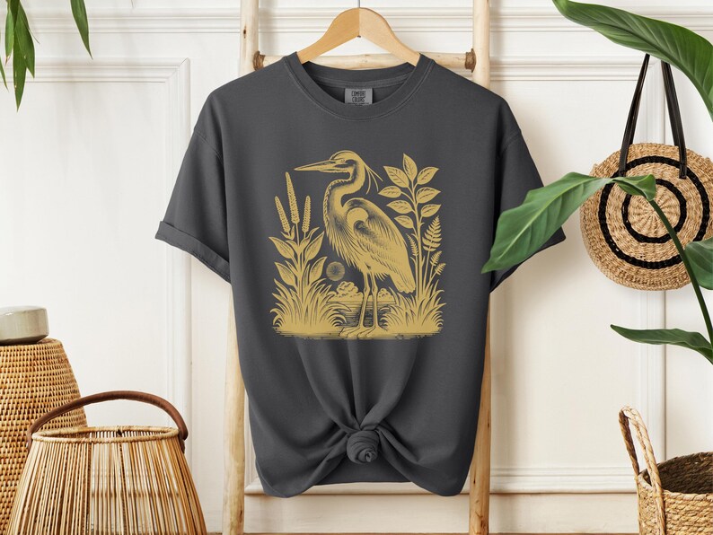 May include: A dark gray t-shirt with a gold illustration of a heron standing in a marsh with reeds and plants.