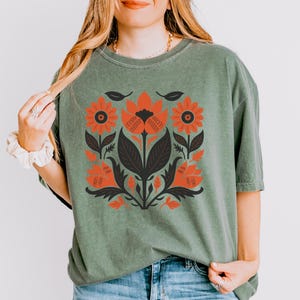 May include: A sage green t-shirt featuring a floral design in shades of orange, black, and dark green. The graphic includes stylized flowers and leaves. The shirt is made of a soft material and has a relaxed fit.