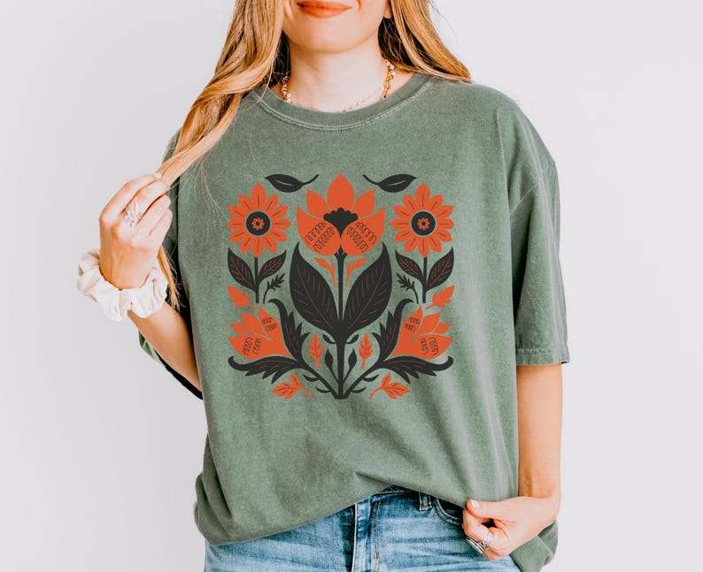 May include: A sage green t-shirt featuring a floral design in shades of orange, black, and dark green. The graphic includes stylized flowers and leaves. The shirt is made of a soft material and has a relaxed fit.