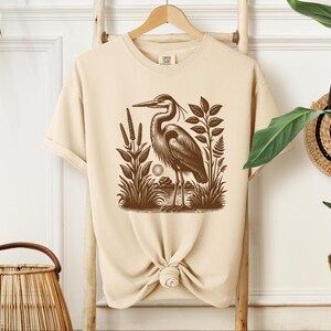 May include: A beige t-shirt with a brown illustration of a heron standing in a marsh. The heron is facing left and has its wings folded. The illustration is surrounded by plants and reeds.