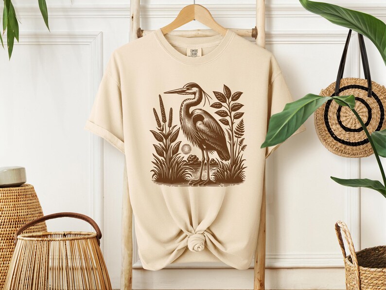 May include: A beige t-shirt with a brown illustration of a heron standing in a marsh. The heron is facing left and has its wings folded. The illustration is surrounded by plants and reeds.
