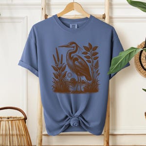 May include: A blue t-shirt with a brown illustration of a heron standing in a marsh. The heron is facing left and has a long neck and beak. The marsh is filled with reeds and grasses.