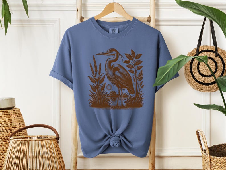 May include: A blue t-shirt with a brown illustration of a heron standing in a marsh. The heron is facing left and has a long neck and beak. The marsh is filled with reeds and grasses.