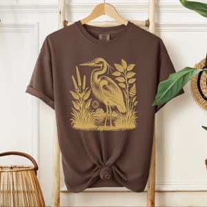 May include: A brown t-shirt with a gold illustration of a heron standing in a marsh. The heron is facing left and has a long neck and beak. The marsh is filled with tall grasses and reeds.