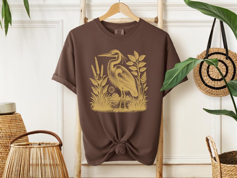 May include: A brown t-shirt with a gold illustration of a heron standing in a marsh. The heron is facing left and has a long neck and beak. The marsh is filled with tall grasses and reeds.