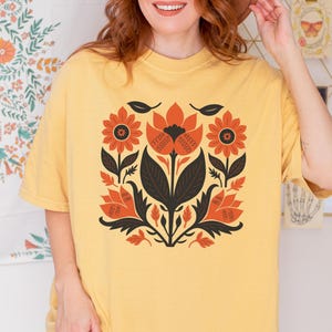 May include: A yellow t-shirt featuring a floral design in orange, black, and white. The design is centered on the shirt and includes stylized flowers and leaves. The shirt has short sleeves and a crew neck.
