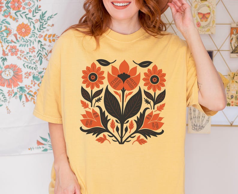 May include: A yellow t-shirt featuring a floral design in orange, black, and white. The design is centered on the shirt and includes stylized flowers and leaves. The shirt has short sleeves and a crew neck.