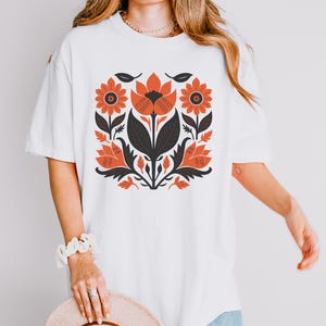 May include: White t-shirt featuring a floral design in shades of orange, black, and dark green. The design includes stylized flowers and leaves, creating a symmetrical pattern. The shirt has short sleeves and a relaxed fit.