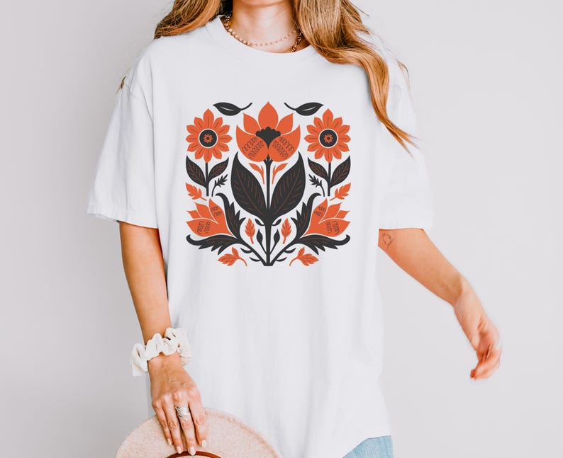 May include: White t-shirt featuring a floral design in shades of orange, black, and dark green. The design includes stylized flowers and leaves, creating a symmetrical pattern. The shirt has short sleeves and a relaxed fit.