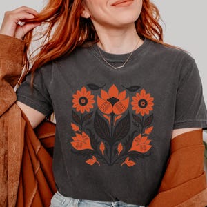 May include: Dark gray t-shirt featuring a symmetrical floral design in orange and black. The design includes stylized flowers, leaves, and stems. The shirt has short sleeves and a crew neck.