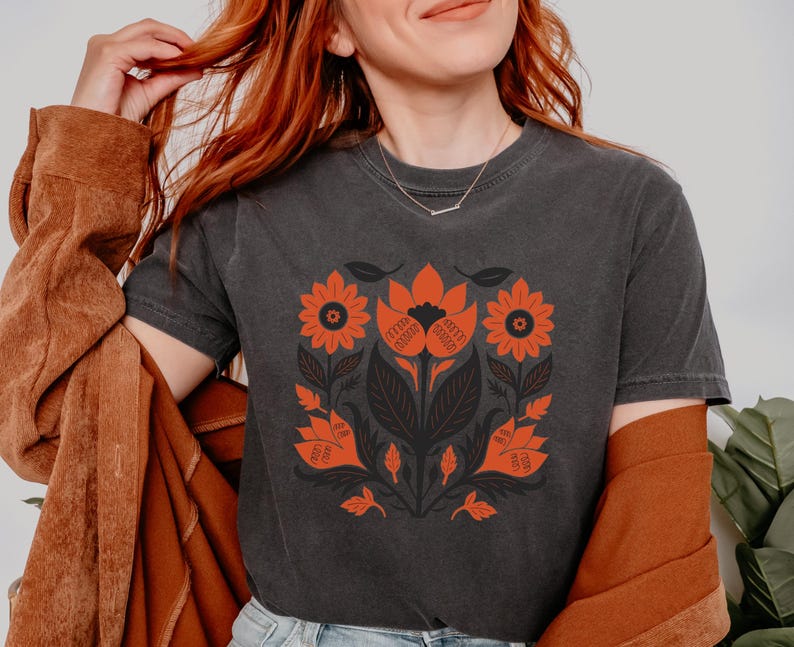 May include: Dark gray t-shirt featuring a symmetrical floral design in orange and black. The design includes stylized flowers, leaves, and stems. The shirt has short sleeves and a crew neck.
