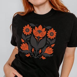 May include: Black t-shirt featuring a floral design in orange and gray. The design includes stylized flowers, leaves, and other botanical elements arranged in a circular pattern. The shirt has a crew neck and short sleeves.