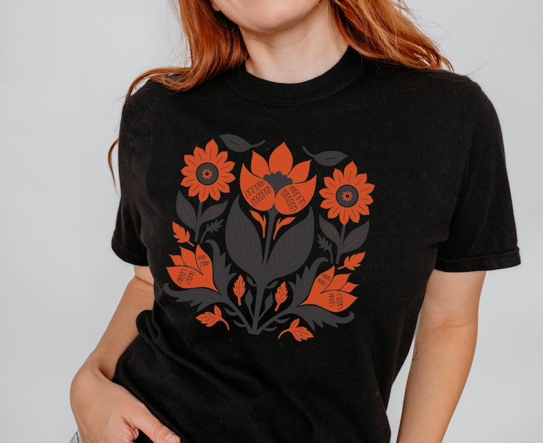 May include: Black t-shirt featuring a floral design in orange and gray. The design includes stylized flowers, leaves, and other botanical elements arranged in a circular pattern. The shirt has a crew neck and short sleeves.