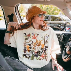 May include: A white t-shirt with a colorful floral print featuring two birds. The print includes leaves, flowers, and berries in shades of orange, yellow, green, and brown.