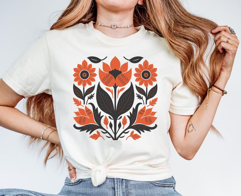 May include: A white t-shirt with a floral design printed on the front. The design features orange and black flowers with a black stem and leaves.