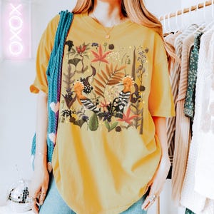 May include: A yellow oversized t-shirt with a colorful floral print featuring two birds. The print includes a variety of flowers, leaves, and branches.