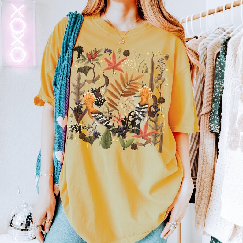 May include: A yellow oversized t-shirt with a colorful floral print featuring two birds. The print includes a variety of flowers, leaves, and branches.