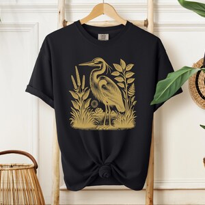 May include: Black t-shirt with a gold illustration of a heron standing in a marsh with reeds and plants.