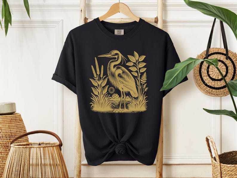 May include: Black t-shirt with a gold illustration of a heron standing in a marsh with reeds and plants.