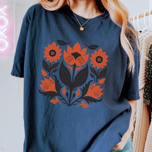May include: Navy blue t-shirt featuring a floral design in orange and black. The design includes stylized flowers, leaves, and stems. The shirt is a comfortable, casual style, suitable for everyday wear.