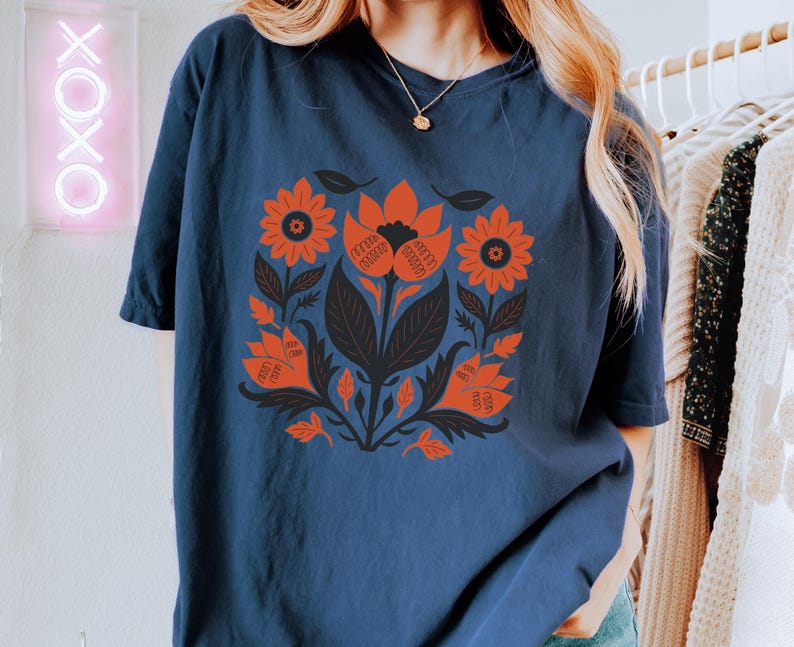 May include: Navy blue t-shirt featuring a floral design in orange and black. The design includes stylized flowers, leaves, and stems. The shirt is a comfortable, casual style, suitable for everyday wear.